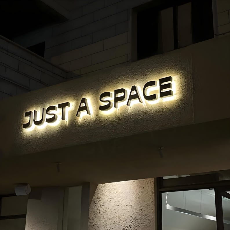 May include: Illuminated sign that reads "JUST A SPACE" in black capital letters. The sign is mounted on a textured, off-white wall, with a warm, yellow glow emanating from behind the letters. The scene is captured at night.