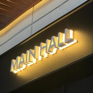 May include: Illuminated sign that reads "MAIN HALL" in white, three-dimensional letters. The sign is mounted on a dark gray wall, with a warm, yellow glow emanating from behind the letters. The sign is angled slightly upward.