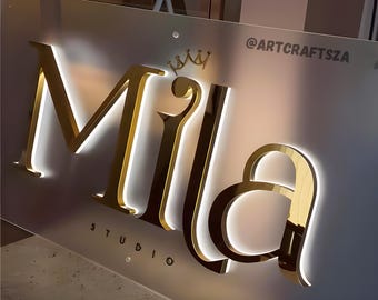 May include: A gold-colored sign with the name "Mila" in large, illuminated letters. The word "STUDIO" is below the name. A small crown graphic is above the name, and the Instagram handle @ARTCRAFTSZA is in the upper right corner.