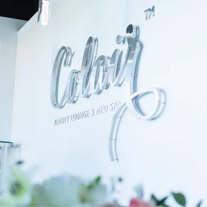 May include: A three-dimensional silver metallic sign mounted on a white wall featuring the cursive text Colair with the words BEAUTY LOUNGE & MED SPA in smaller block letters underneath and a small TM symbol in the upper right corner.