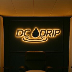May include: A wall-mounted sign featuring the text DC DRIP in bold, capitalized letters with a central water drop icon and concentric ripple lines below, all illuminated by a warm, golden backlighting against a dark, matte-finished wall.