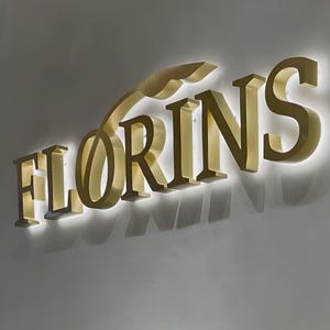 May include: A three-dimensional wall sign featuring the word FLORINS in large, metallic gold capital letters. The letters have a brushed finish and are mounted with a backlighting effect that casts a soft white glow onto the wall behind them.