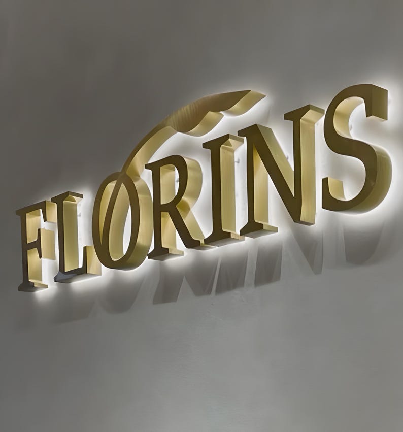 May include: A three-dimensional wall sign featuring the word FLORINS in large, metallic gold capital letters. The letters have a brushed finish and are mounted with a backlighting effect that casts a soft white glow onto the wall behind them.
