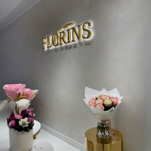 May include: A wall-mounted gold 3D sign reading FLORINS with backlighting, positioned above two floral arrangements. One bouquet is in a white cylindrical vase, and the other is in a glass vase on a gold pedestal, featuring pink and white roses.