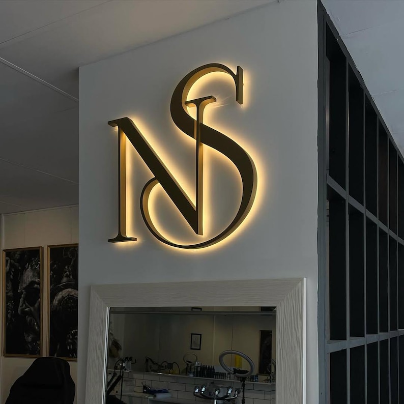May include: A gold-colored illuminated sign displaying the intertwined letters "NS" mounted on a white wall. The sign is lit from behind, creating a warm glow. The sign is above a mirror and below a dark metal structure.