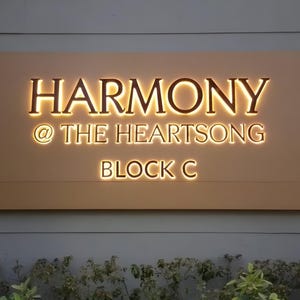 May include: A rectangular sign with the words "HARMONY @ THE HEARTSONG BLOCK C" in illuminated, warm-toned lettering. The sign is mounted on a light-colored surface, with a background of a gray wall and some greenery.