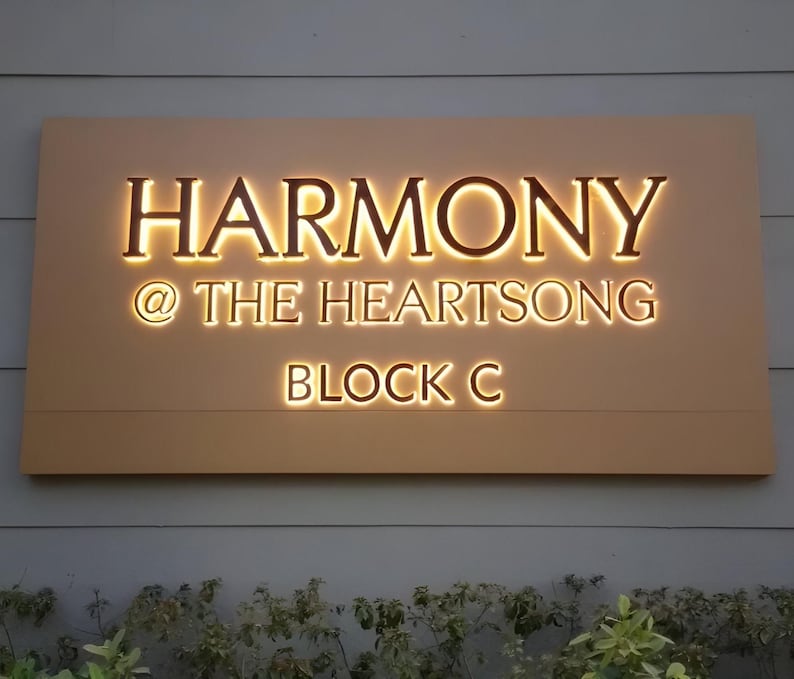 May include: A rectangular sign with the words "HARMONY @ THE HEARTSONG BLOCK C" in illuminated, warm-toned lettering. The sign is mounted on a light-colored surface, with a background of a gray wall and some greenery.