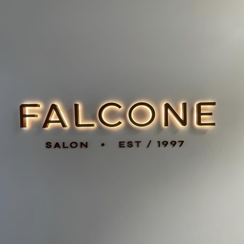 May include: A wall-mounted sign for FALCONE SALON, with the words illuminated in a warm, golden light. Below, the text reads "SALON &bull; EST / 1997" in a smaller font. The sign is mounted on a neutral-colored wall.