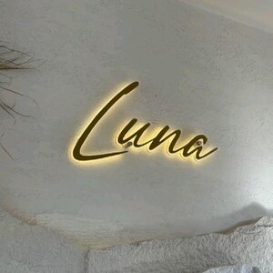 May include: A wall-mounted sign featuring the word Luna in a gold-toned, cursive script font. The letters are backlit with a warm, glowing light that creates a soft halo effect against the textured, off-white surface behind the lettering.