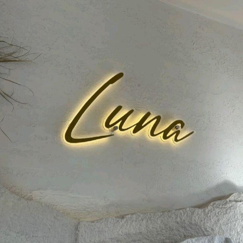May include: A wall-mounted sign featuring the word Luna in a gold-toned, cursive script font. The letters are backlit with a warm, glowing light that creates a soft halo effect against the textured, off-white surface behind the lettering.