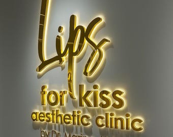 May include: A gold-colored illuminated sign on a wall. The sign reads "lips" in a stylized script, followed by "for kiss aesthetic clinic" and "by Dr. Marina Aisina" in a smaller font. The sign is lit from behind, creating a glowing effect.