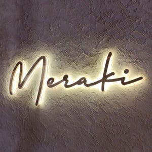 May include: A wall-mounted sign featuring the word Meraki written in a cursive script font. The letters are dark in color and are illuminated from behind by a warm white light, creating a glowing halo effect against the textured wall surface.