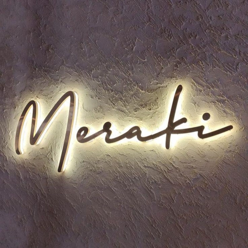 May include: A wall-mounted sign featuring the word Meraki written in a cursive script font. The letters are dark in color and are illuminated from behind by a warm white light, creating a glowing halo effect against the textured wall surface.