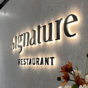 May include: A wall-mounted sign featuring the word signature in lowercase black letters above the word RESTAURANT in uppercase black letters, both illuminated from behind with a warm glow against a light gray textured wall surface.