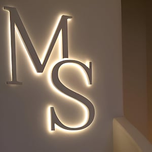 May include: Illuminated white metal letters 'M' and 'S' mounted on a wall. The letters have a warm, glowing light outlining their edges, creating a striking visual effect. The design is modern and minimalist.
