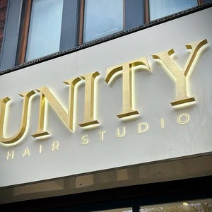 以下が含まれることがあります： 「UNITY HAIR STUDIO」の看板。金色の立体的な文字で表示されています。白い表面に取り付けられており、下には小さな金色の文字で「HAIR STUDIO」と書かれています。背景には窓が見えます。