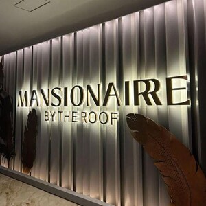 May include: A wall display featuring the gold-colored text MANSIONAIRE BY THE ROOF mounted on a vertical panel background with integrated backlighting. Two large, brown, textured feather-shaped decorative elements flank the lettering.