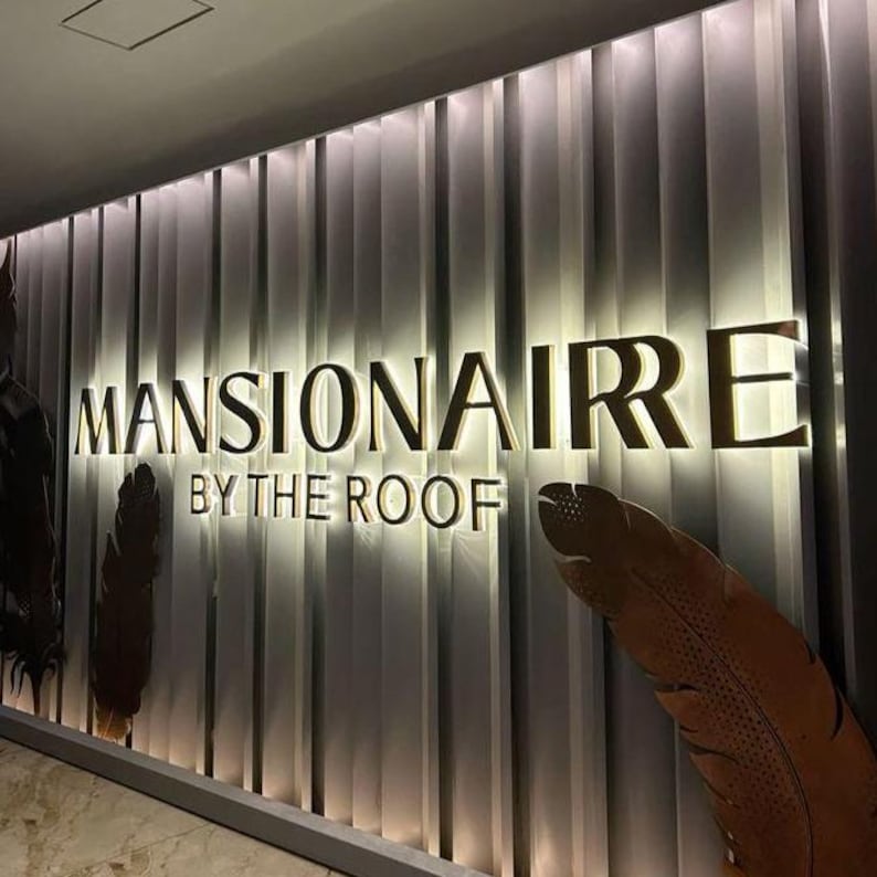 May include: A wall display featuring the gold-colored text MANSIONAIRE BY THE ROOF mounted on a vertical panel background with integrated backlighting. Two large, brown, textured feather-shaped decorative elements flank the lettering.