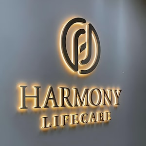 以下が含まれることがあります： 「HARMONY LIFECARE」の文字がセリフ体で書かれた照明付きの看板。テキストの上には葉のデザインの円形ロゴがあります。看板は灰色の壁に取り付けられており、暖かく金色の光を放っています。