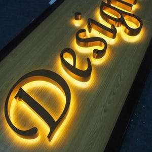 May include: A three-dimensional sign featuring the word Design in gold-colored metal letters mounted on a light wood-grain surface. Each letter is backlit with a warm yellow glow, creating a halo effect that illuminates the wood texture.
