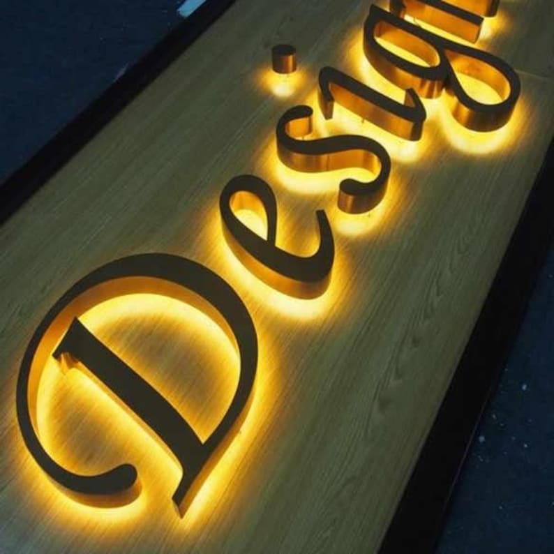 May include: A three-dimensional sign featuring the word Design in gold-colored metal letters mounted on a light wood-grain surface. Each letter is backlit with a warm yellow glow, creating a halo effect that illuminates the wood texture.