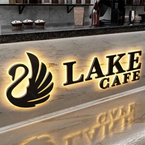 May include: A backlit sign on a marble counter featuring a black swan silhouette logo and the text LAKE CAFE in bold, dark, raised lettering. The sign is illuminated from behind, casting a warm glow against the white and grey marble surface.