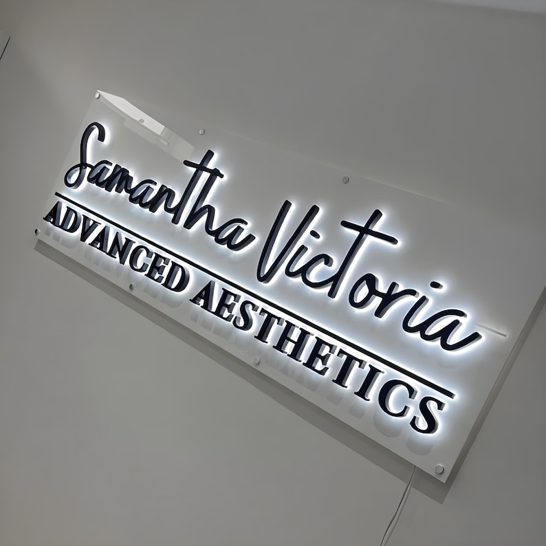 May include: A rectangular wall-mounted sign featuring the black text Samantha Victoria in a script font and ADVANCED AESTHETICS in a bold sans-serif font. The sign has a white acrylic backing with integrated LED backlighting behind the letters.