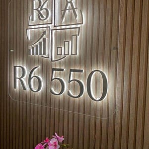 May include: A rectangular clear acrylic sign featuring a backlit logo with the text R6 550. The logo includes a compass, a rising bar graph, and the letters R6. The sign is mounted on a vertical wooden slat wall, with pink orchid flowers visible below.