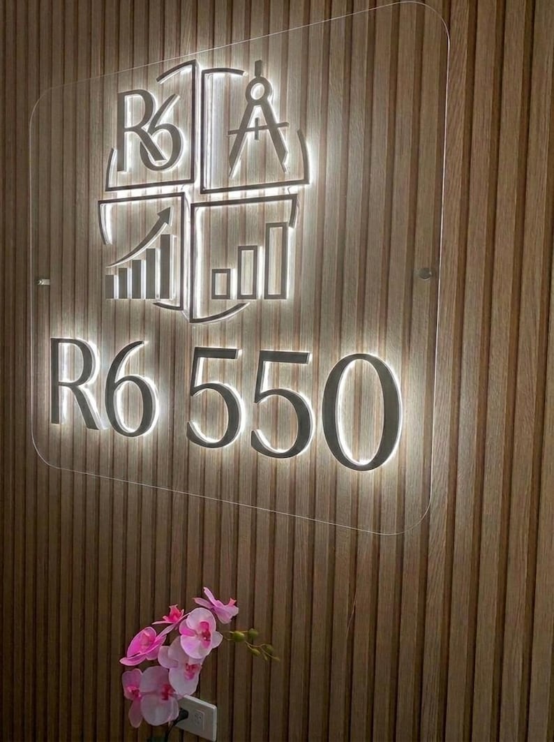 May include: A rectangular clear acrylic sign featuring a backlit logo with the text R6 550. The logo includes a compass, a rising bar graph, and the letters R6. The sign is mounted on a vertical wooden slat wall, with pink orchid flowers visible below.