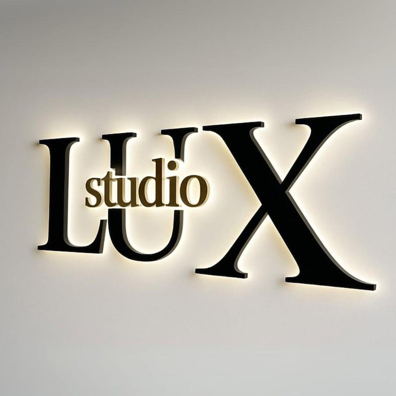 May include: A wall-mounted sign featuring the large black letters L, U, and X with a smaller gold-colored word studio positioned between the U and X. The letters are backlit, creating a soft glowing halo effect against the plain wall surface.