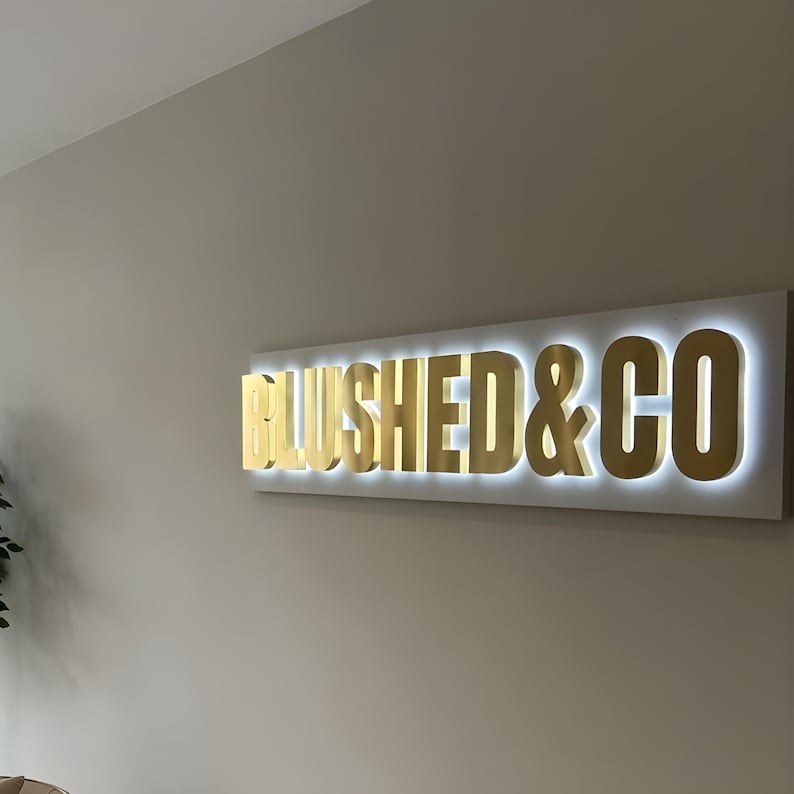 May include: A rectangular wall sign featuring the gold-colored, three-dimensional text BLUSHED & CO mounted on a white rectangular backing. The letters are illuminated from behind with a bright white light, creating a glowing halo effect.