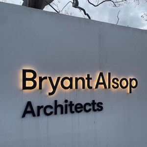 May include: A white wall featuring black dimensional lettering that reads Bryant Alsop Architects. The top line, Bryant Alsop, is illuminated from behind with a warm glow, while the word Architects below is in solid black lettering without lighting.