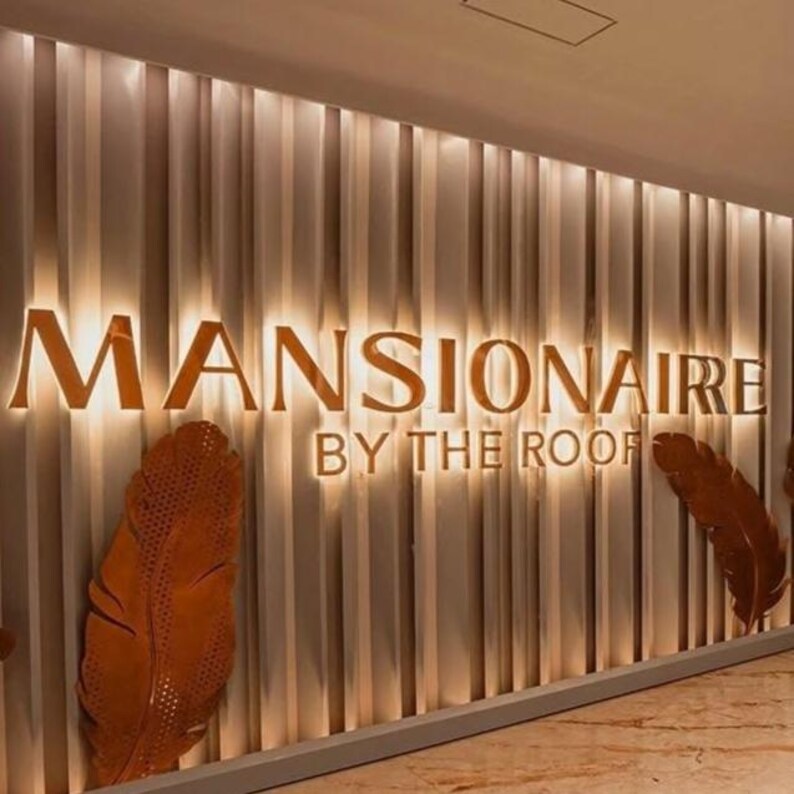 May include: A wall display featuring the gold-colored text MANSIONAIRE BY THE ROOF mounted on a vertical paneled background with integrated warm lighting. Two large, textured, copper-toned metal feathers are positioned on the lower sides.