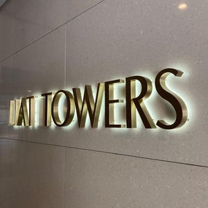 May include: A wall-mounted sign featuring three-dimensional gold-toned metal letters that spell out "LAT TOWERS". The letters are backlit with a soft white glow, creating a halo effect against the smooth, light beige speckled stone wall surface.