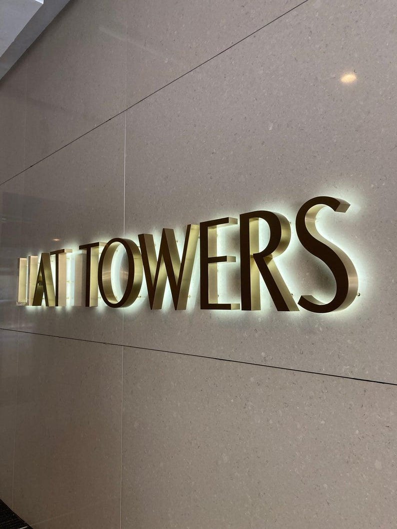 May include: A wall-mounted sign featuring three-dimensional gold-toned metal letters that spell out "LAT TOWERS". The letters are backlit with a soft white glow, creating a halo effect against the smooth, light beige speckled stone wall surface.