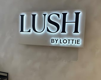 May include: A rectangular wall-mounted sign with a white background and black lettering that reads LUSH BY LOTTIE. The letters are backlit with a bright white glow, creating a halo effect around the edges of the text against the white panel.