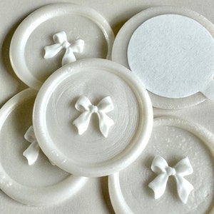 May include: Five white round wax seals with a raised white bow design. The seals are arranged in a circle with one seal in the center.