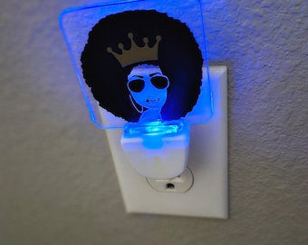 May include: A white night light with a blue glow. The night light has a clear plastic insert with a black and white image of a woman with an afro wearing sunglasses and a gold crown.