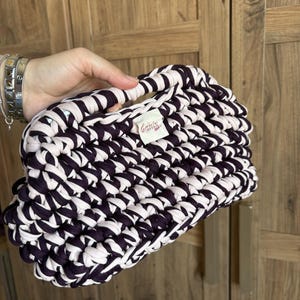 May include: A handmade crochet handbag crafted from thick, dark purple and light pink fabric yarn. The bag features a textured, chunky knit pattern with an integrated handle and a small white fabric label with the text Canix printed in red.