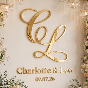 May include: A large gold-colored reflective monogram featuring the letters C and L in a cursive script, positioned above the text Charlotte & Leo and the date 07.07.26, all mounted on a wall flanked by vertical arrangements of white and peach roses.