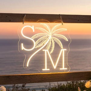 Puede incluir: Una escena de boda en la playa con una valla de madera adornada con rosas blancas y un letrero de neón con las letras "S" y "M" y una palmera. La escena está ambientada en una playa de arena con conchas marinas y madera flotante.