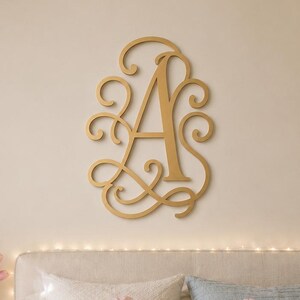 May include: A gold-coloured wall-mounted letter A monogram featuring an ornate, swirling script design. The letter is displayed above a bed with light-coloured bedding, including a pink gingham pillow and a white pillow with the embroidered letters AMG.