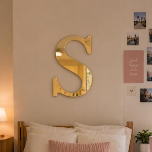 Op de afbeelding: Een slaapkamerinterieur met een grote goudkleurige letter S aan de muur boven een bed met wit beddengoed, een roze textuurrijke sprei en een bijpassend roze lendenkussen. Een witte rugzak staat op de vloer bij een houten ladekast.