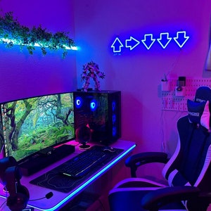 May include: A gaming setup with a curved monitor displaying a forest scene. The desk has a keyboard, mouse, and headset. A neon blue arrow sign hangs on the wall. A gaming chair and a computer tower with blue lights complete the setup.