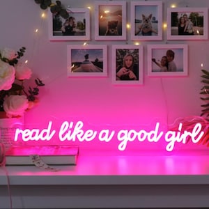 Puede incluir: Un letrero de neón rosa brillante con la frase "read like a good girl" en escritura cursiva. El letrero está montado en una pared rosa, debajo de fotos enmarcadas y luces de hadas. Un ramo de flores blancas y libros están en un estante blanco.