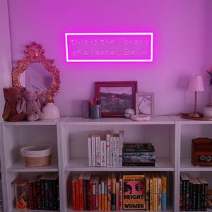 May include: A neon sign with the text "this is the library of a reader. Bella" hangs above a white bookshelf filled with books. A gold-framed mirror and decorative items sit on the shelf. The room is lit with a pink glow.