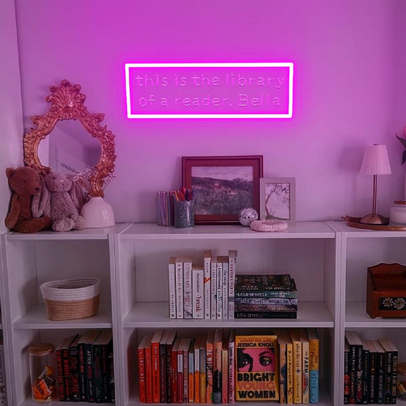 May include: A neon sign with the text "this is the library of a reader. Bella" hangs above a white bookshelf filled with books. A gold-framed mirror and decorative items sit on the shelf. The room is lit with a pink glow.