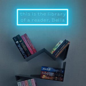 May include: A modern bookshelf with several books, including titles like "Nightbane" and "Mile High." A neon sign above reads "this is the library of a reader, Bella." The shelves are gray and mounted on a light blue wall.