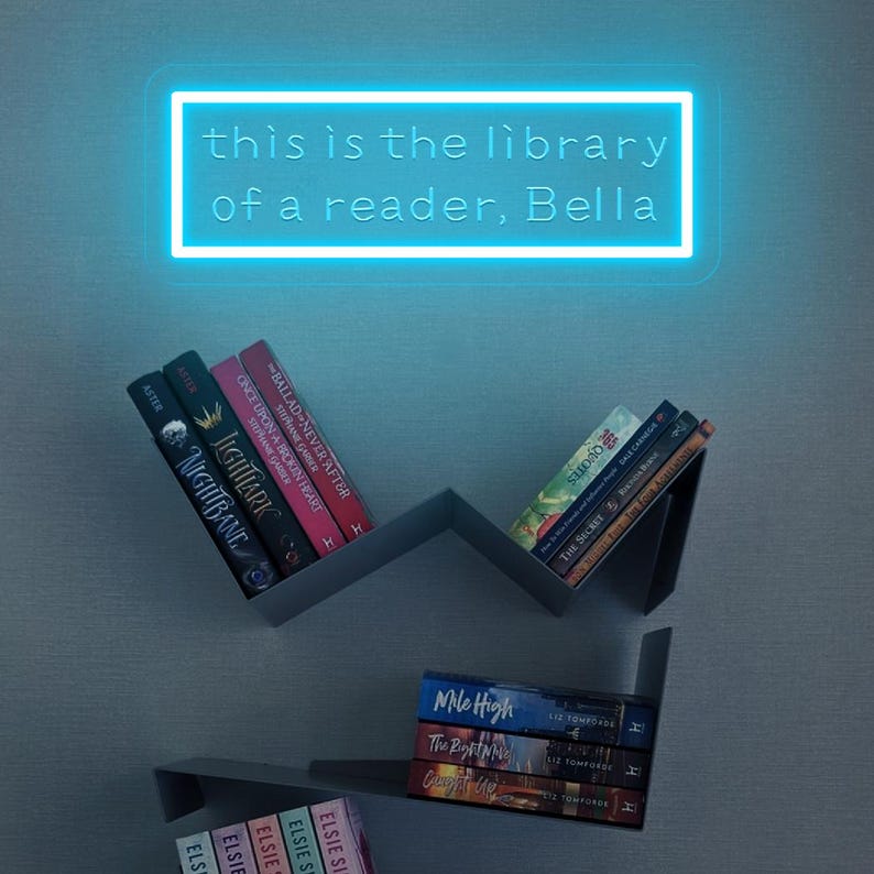 May include: A modern bookshelf with several books, including titles like "Nightbane" and "Mile High." A neon sign above reads "this is the library of a reader, Bella." The shelves are gray and mounted on a light blue wall.