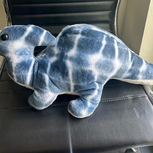 May include: A plush dinosaur toy featuring a soft, fuzzy texture with a blue and white tie-dye pattern. The stuffed animal has a prominent crest on its head, a long tail, four short legs, and a single visible black button eye on its side.